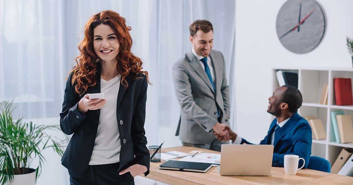 Eine junge Frau steht in einem B&uuml;ro und ist gl&uuml;cklich &uuml;ber einen neuen Job, im Hintergrund sch&uuml;tteln sich zwei M&auml;nner die Hand | Immobilienverkauf bei Jobwechsel