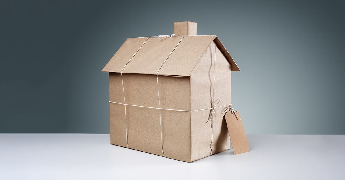 Ein Modell eines Hauses steht in braunem Papier verpackt und mit Schnur zusammengebunden auf einem wei&szlig;en Tisch vor einem dunklen Hintergrund | Immobilie erben